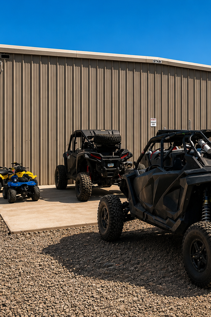 A black off-road side-by-side UTV with a roll cage and large knobby tires parked on gravel in front of a tan metal building, with other ATVs and a red UTV nearby under a clear blue sky.